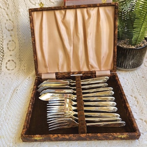 以下が含まれることがあります： 木製の箱に入った、オープンなヴィンテージのカトラリーセット。フォーク、ナイフ、スプーンが含まれており、すべて装飾的なハンドルが付いています。箱は茶色で、装飾的な模様と金色の留め金が付いています。