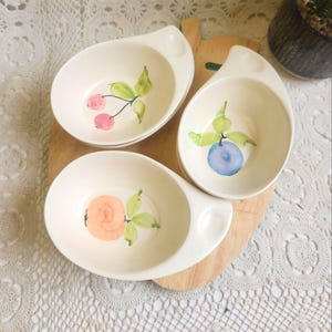May include: Three off-white ceramic bowls with a handle, each decorated with a different hand-painted fruit design: cherries, a blueberry, and a peach. The bowls are arranged on a wooden cutting board.