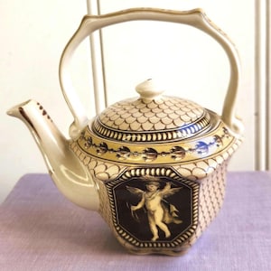 May include: Cream-colored ceramic teapot with a decorative handle and spout. The teapot features a detailed design with a cherub illustration on the front, and a patterned lid. The teapot has a yellow and brown border.