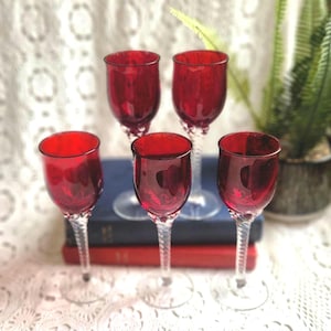 May include: A set of four ruby red glass goblets with clear, twisted stems. Each glass features a rounded bowl and a circular base. The glasses are arranged in a group, with two positioned in the back and two in the front, showcasing the deep red color and the intricate spiral pattern of the glass stems.