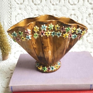 May include: A brown ceramic vase with a scalloped rim and a wood grain texture. The exterior is decorated with small, raised, multi-coloured floral appliques in shades of yellow, blue, pink, and green, arranged along the top edge and base.