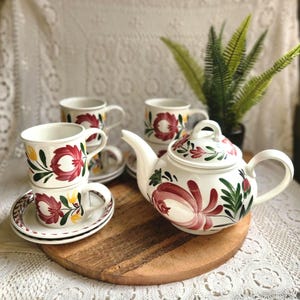 May include: A white ceramic tea set featuring a hand-painted floral pattern in shades of red, green, and yellow. The set includes a teapot with a lid and multiple matching cups and saucers, all decorated with stylised botanical motifs.