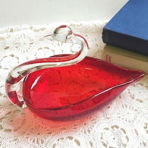 May include: A red glass swan-shaped dish with a clear glass neck and head. The dish has a textured interior and sits on a white lace surface. A blue book is visible in the background.