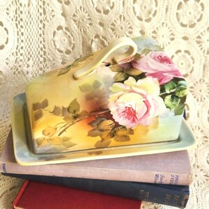 May include: A ceramic butter dish with a rectangular base and a domed lid featuring a handle. The surface is decorated with a hand-painted floral pattern of large pink roses, green leaves, and soft yellow and blue watercolor-style shading.