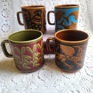 May include: Four ceramic mugs with distinctive designs. One mug features a rooster, another dolphins, a third rabbits, and the last a mythical creature. The mugs have brown, green, red, and blue backgrounds with various colours for the designs.