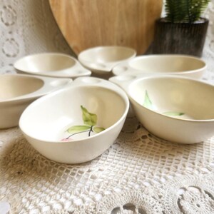 May include: A collection of off-white ceramic bowls, some with attached compartments. Each bowl features a hand-painted design of a green leaf and a pink cherry. The bowls are arranged on a white lace tablecloth, suggesting a vintage or retro aesthetic.