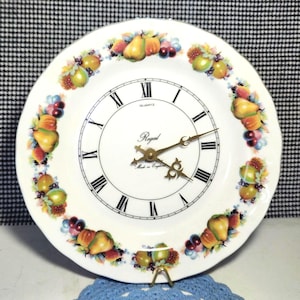 May include: A white ceramic clock featuring a fruit design around the edge. The clock face has black Roman numerals and gold hands. The word "Regal" is printed on the clock face, along with "Quartz" and "Made in England".