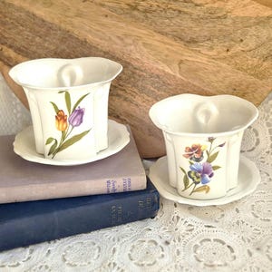 May include: Two white porcelain cups with matching saucers, each featuring a scalloped rim and a central thumb rest. One cup displays a painted orange and purple tulip, while the other features a multi-coloured pansy floral illustration.