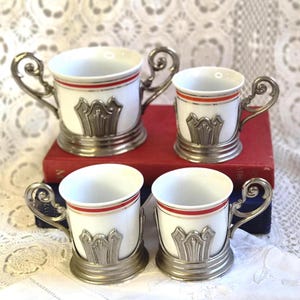 May include: A set of four white porcelain cups with red and silver horizontal stripes, each encased in a decorative silver-toned metal holder featuring ornate scroll handles and a raised architectural emblem on the front base.