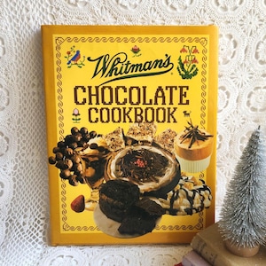 May include: A vintage "Whitman's Chocolate Cookbook" with a yellow cover. The cover features the title in large, pixelated letters, along with illustrations of chocolate desserts. A small, silver Christmas tree is visible in the background.