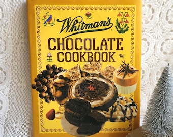 May include: A vintage "Whitman's Chocolate Cookbook" with a yellow cover. The cover features the title in large, pixelated letters, along with illustrations of chocolate desserts. A small, silver Christmas tree is visible in the background.