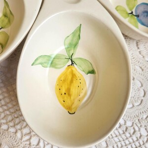 May include: Cream-colored ceramic serving dish with a hand-painted yellow lemon and green leaves. The lemon has black speckles and is centered in the oval-shaped bowl. The dish has a handle and is part of a set.