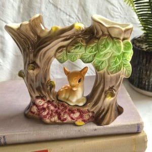 May include: A ceramic vase shaped like a tree trunk with a glossy brown finish, featuring sculpted green leaves at the top, pink flowers at the base, and a small, light brown fawn with white spots resting in the centre opening of the tree structure.