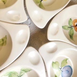 May include: A collection of off-white ceramic serving dishes. Each dish has a unique shape with a curved edge and a small, round indentation. The dishes are decorated with hand-painted fruit and leaf designs in shades of green, orange, and blue.