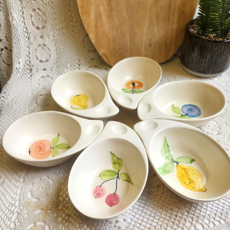 May include: Six off-white ceramic serving bowls with a unique handle design. Each bowl features a different hand-painted fruit or floral design, including a pear, orange flower, blueberry, rose, cherries, and a lemon. The bowls are arranged on a white lace tablecloth.