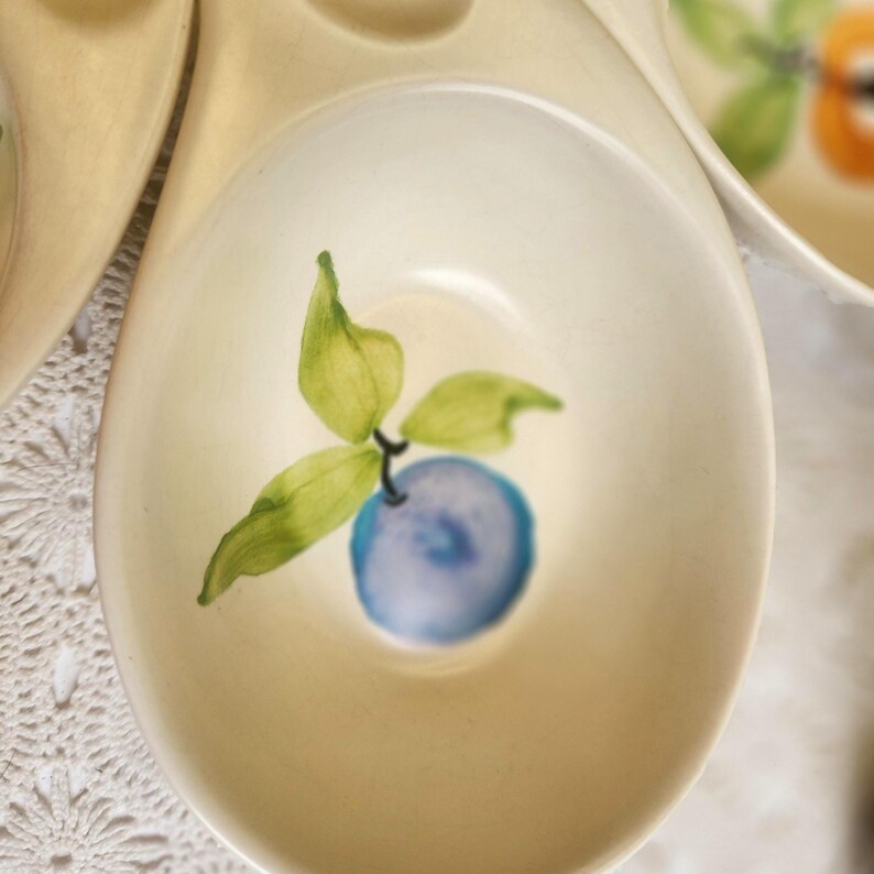 May include: Cream-colored ceramic serving dish with a hand-painted design of a blue fruit and green leaves. The dish has a shallow, rounded bowl shape and a curved handle. The fruit design is centered in the bowl.