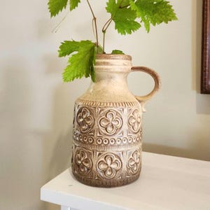 May include: A beige ceramic vase with a handle and a floral pattern. The vase has a textured surface and contains green leaves. The vase is on a white surface. The vase is approximately 20 cm tall.