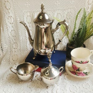 May include: A silver-plated tea set comprising a tall teapot with a lid, a creamer, and a sugar bowl, all featuring ornate feet. Beside these are two white porcelain teacups with pink rose patterns on saucers, arranged on a dark blue book.