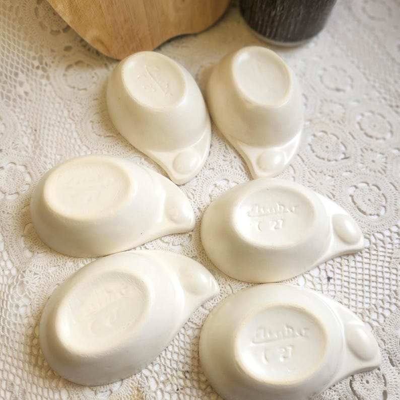 May include: A collection of five off-white ceramic dishes. Each dish has a teardrop shape with a small, rounded handle and a slightly raised rim. The bottoms of the dishes are stamped with the word "Dunbar" and the number "C27".