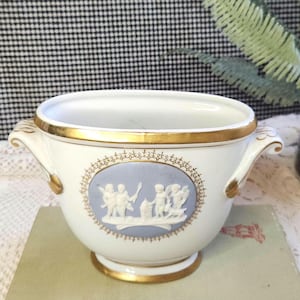 May include: A white ceramic bowl with gold trim and handles. The bowl features a light blue oval medallion with a classical relief scene. The rim and base are also trimmed in gold, creating an elegant, vintage aesthetic.