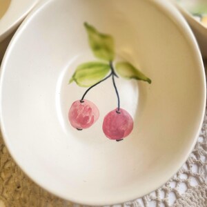 May include: A small, off-white ceramic bowl with a hand-painted cherry design. The cherries are pink with black stems and green leaves. The bowl has a shallow, rounded shape, suitable for serving small portions of food.