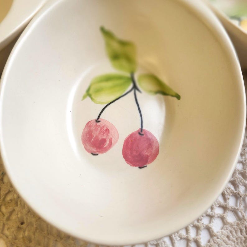 May include: A small, off-white ceramic bowl with a hand-painted cherry design. The cherries are pink with black stems and green leaves. The bowl has a shallow, rounded shape, suitable for serving small portions of food.