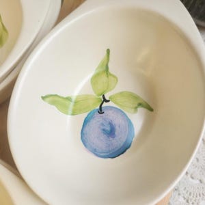 May include: Off-white oval serving dish with a hand-painted design of a blue fruit with green leaves. The dish has a smooth, glossy finish and a slightly raised edge. The fruit design is centered in the dish.