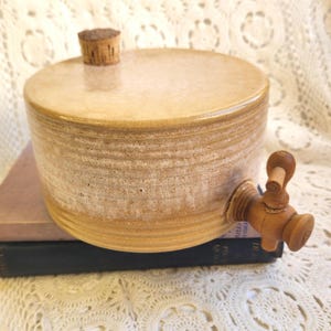 May include: A cylindrical tan ceramic beverage dispenser with a smooth, glazed finish and horizontal ridged detailing. The vessel features a small cork stopper on the top surface and a wooden spigot attached to the side for dispensing liquids.