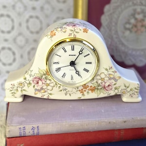 May include: A cream-colored ceramic mantel clock with a gold-tone bezel and a white face featuring black Roman numerals and the text "STAIGER" and "QUARTZ". The clock body is decorated with a floral pattern of pink roses and orange flowers.