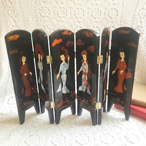 May include: A small, folding room divider with a black lacquered finish. The divider features painted panels depicting figures in traditional attire, with accents of red, orange, and white. The panels are connected by brass hinges.