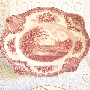 May include: An oval ceramic platter featuring a deep red transferware pattern on a cream background. The center depicts a riverside landscape with a bridge, buildings, and boats. The border is decorated with intricate floral and lattice motifs.