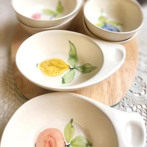 May include: A collection of off-white ceramic bowls with hand-painted fruit designs. One bowl features a yellow lemon with green leaves, while others display pink and blue fruit. The bowls are arranged on a wooden surface.