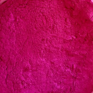 Pode incluir: Close-up de um p&oacute; f&uacute;csia vibrante com uma superf&iacute;cie texturizada. O p&oacute; parece finamente mo&iacute;do e uniformemente distribu&iacute;do, sugerindo um produto cosm&eacute;tico ou alimentar. A cor &eacute; consistente em toda a imagem.