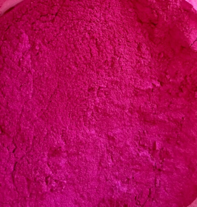 Pode incluir: Close-up de um p&oacute; f&uacute;csia vibrante com uma superf&iacute;cie texturizada. O p&oacute; parece finamente mo&iacute;do e uniformemente distribu&iacute;do, sugerindo um produto cosm&eacute;tico ou alimentar. A cor &eacute; consistente em toda a imagem.