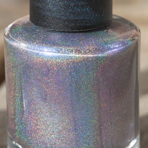 May include: A clear glass nail polish bottle with a black cap. The polish inside is a shimmering, iridescent color that shifts between purple, silver, and green. The bottle is cylindrical and sits on a wooden surface.