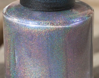 Puede incluir: Un frasco de esmalte de uñas de vidrio transparente con tapa negra. El esmalte tiene un efecto brillante e iridiscente, que cambia entre púrpura, plateado y verde. El frasco cilíndrico está sobre una superficie de madera.