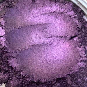 May include: Close-up of a container filled with a shimmering, metallic purple powder. The powder has a smooth, textured appearance with visible brush strokes, creating a gradient of light and shadow. The container's rim is visible.