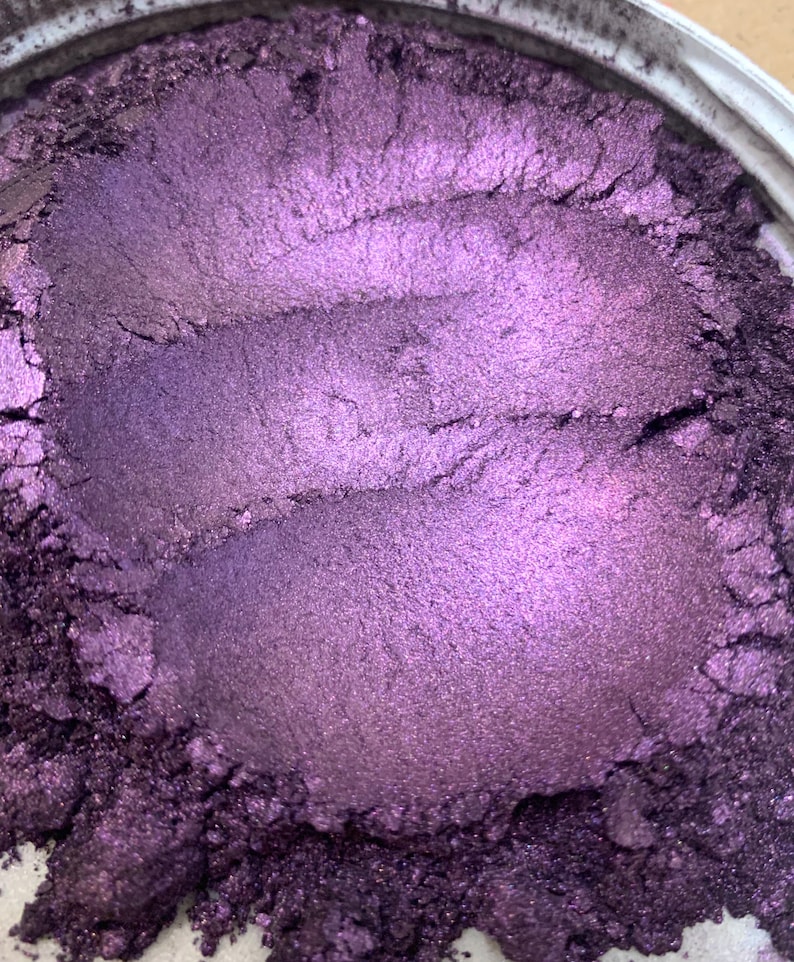 May include: Close-up of a container filled with a shimmering, metallic purple powder. The powder has a smooth, textured appearance with visible brush strokes, creating a gradient of light and shadow. The container's rim is visible.