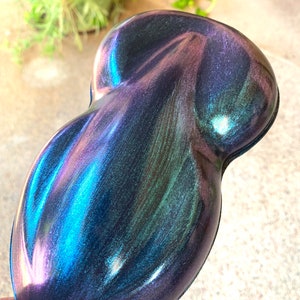 May include: An iridescent, teardrop-shaped object with a smooth, glossy surface. The color shifts from deep blues and purples to hints of green and pink, creating a dynamic, shimmering effect. The object's form is rounded and ergonomic.