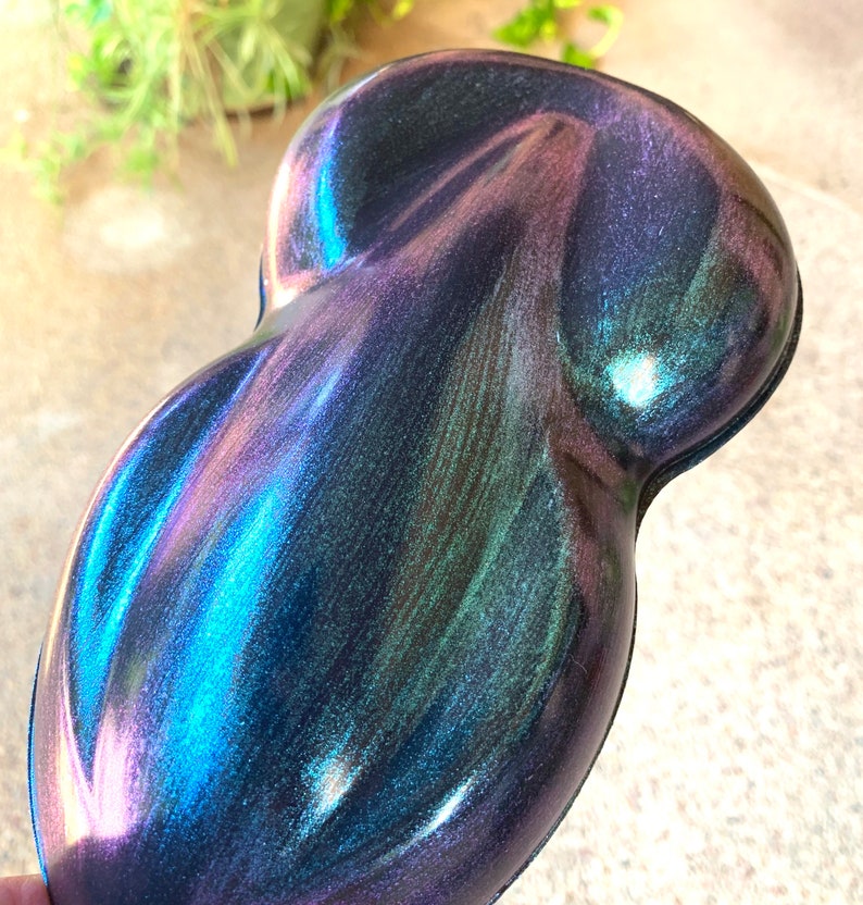 May include: An iridescent, teardrop-shaped object with a smooth, glossy surface. The color shifts from deep blues and purples to hints of green and pink, creating a dynamic, shimmering effect. The object's form is rounded and ergonomic.