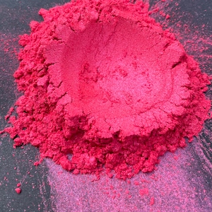 May include: A close-up of a pile of vibrant, finely milled, pink pigment powder. The powder has a metallic sheen and a textured appearance, with small particles visible. The color is a bright, saturated pink, with variations in tone.