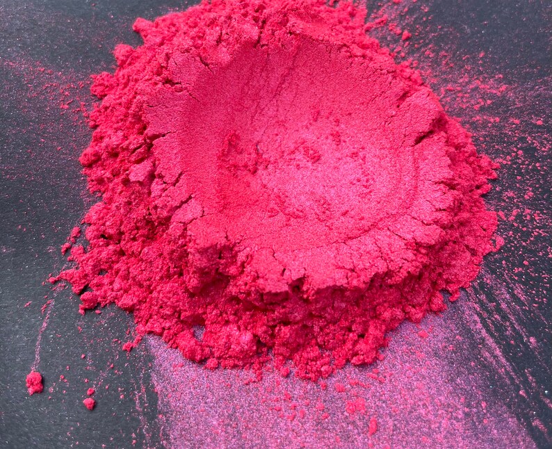 May include: A close-up of a pile of vibrant, finely milled, pink pigment powder. The powder has a metallic sheen and a textured appearance, with small particles visible. The color is a bright, saturated pink, with variations in tone.
