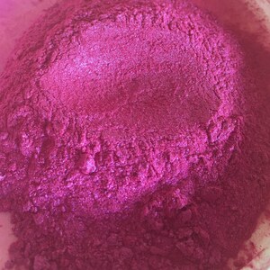 May include: Close-up of a pile of vibrant, magenta-colored powder. The powder has a shimmering, metallic sheen, suggesting a pigment or cosmetic product. The texture appears fine and granular, with subtle variations in tone.