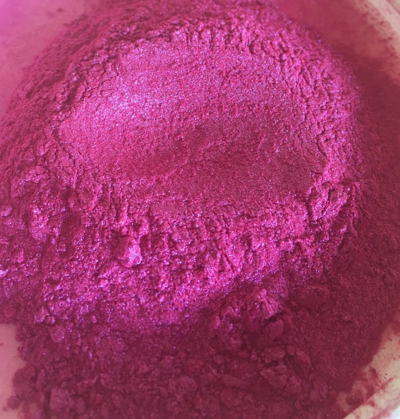 May include: Close-up of a pile of vibrant, magenta-colored powder. The powder has a shimmering, metallic sheen, suggesting a pigment or cosmetic product. The texture appears fine and granular, with subtle variations in tone.