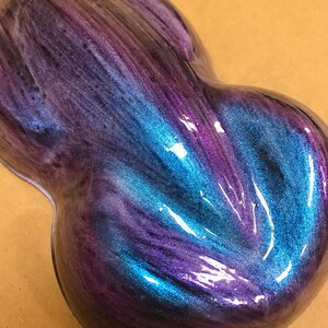 May include: A glossy, abstract sculpture with a flowing design. The piece features a blend of iridescent colours, including deep purples, blues, and hints of teal. The surface reflects light, creating a dynamic visual effect.