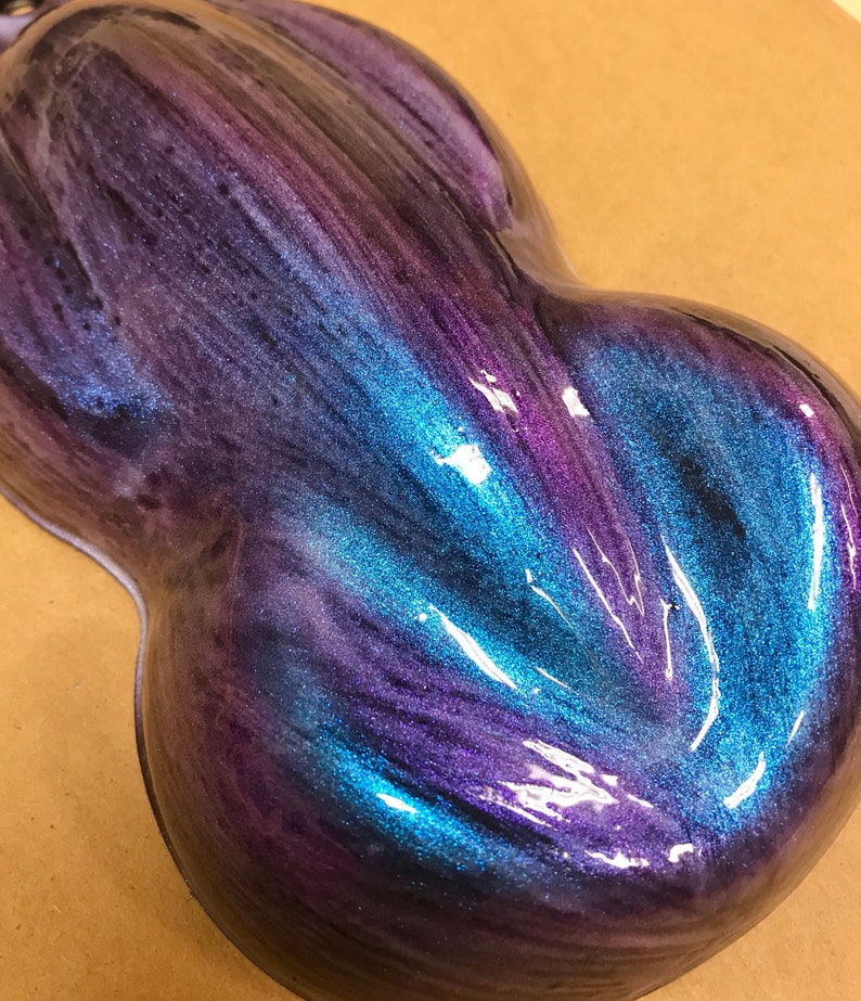 May include: A glossy, abstract sculpture with a flowing design. The piece features a blend of iridescent colours, including deep purples, blues, and hints of teal. The surface reflects light, creating a dynamic visual effect.