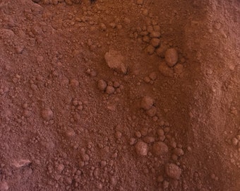 May include: Close-up of a pile of fine, reddish-brown powder with a slightly coarse texture. The powder has small, irregular clumps scattered throughout, creating a varied surface. The color is consistent, with subtle variations in tone.
