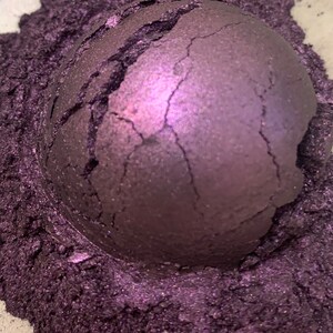 May include: A close-up of a cracked, spherical object in a deep purple hue, surrounded by a bed of fine, shimmering purple powder. The sphere's surface has a textured appearance with visible cracks.