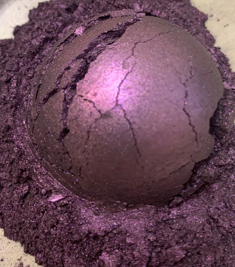 May include: A close-up of a cracked, spherical object in a deep purple hue, surrounded by a bed of fine, shimmering purple powder. The sphere's surface has a textured appearance with visible cracks.