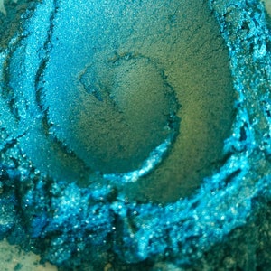 May include: Close-up of a pile of teal-colored pigment powder. The powder has a shimmering, metallic finish and a swirling pattern in the center. The texture appears fine and granular, with variations in color and depth.